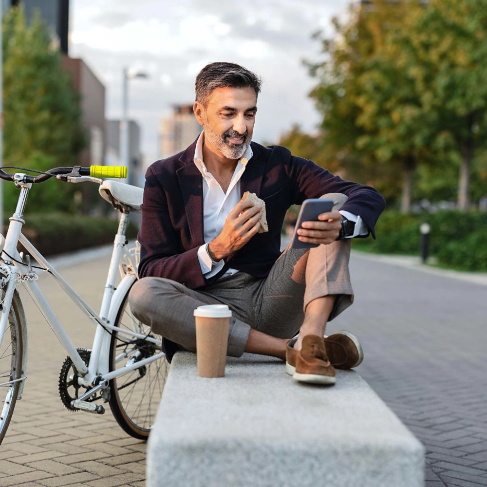 Mann sitzt auf einer Bank, schaut auf sein Handy und hält einen Kaffee, neben einem Fahrrad.