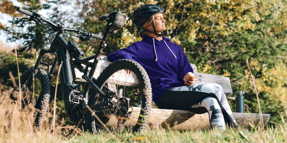 Frau in lila Hoodie entspannt auf einer Bank neben einem E-Bike im Freien
